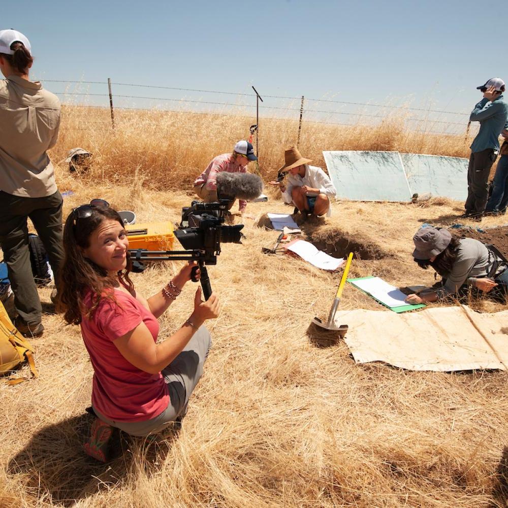 当土壤科学学生在干秸秆景观中取样土壤时，穿着红衬衫的女子带着摄像机微笑